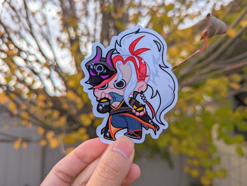 May include: A sticker featuring a cartoon character with white hair, a red and black outfit, and a black mask. The character is holding a weapon in their right hand and has a determined expression.