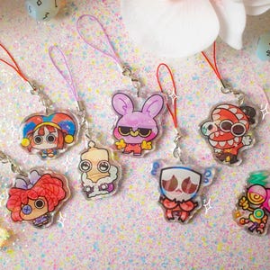 May include: A collection of seven acrylic charms featuring colorful cartoon characters, each attached to a thin nylon cord loop. The designs include a jester, a rabbit, a creature with large eyes, a robot, and other stylized figures with vibrant details.
