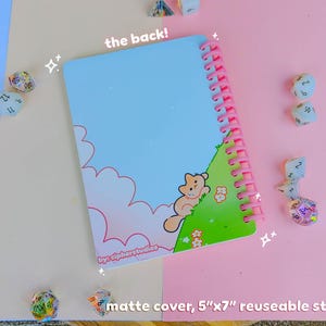 May include: A matte cover, 12.7cm x 17.8cm reusable sticker book with a light blue sky design and a pink spiral binding. The cover features a cartoon cat on a green hill. The text "the back!" is visible.