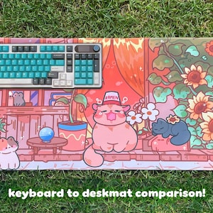 May include: A colorful desk mat featuring a cartoon cat design with sunflowers and other plants. A teal and white keyboard sits on the mat. The text "keyboard to deskmat comparison!" is at the bottom.
