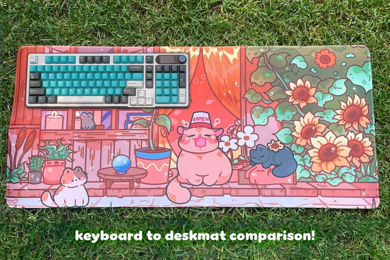May include: A colorful desk mat featuring a cartoon cat design with sunflowers and other plants. A teal and white keyboard sits on the mat. The text "keyboard to deskmat comparison!" is at the bottom.