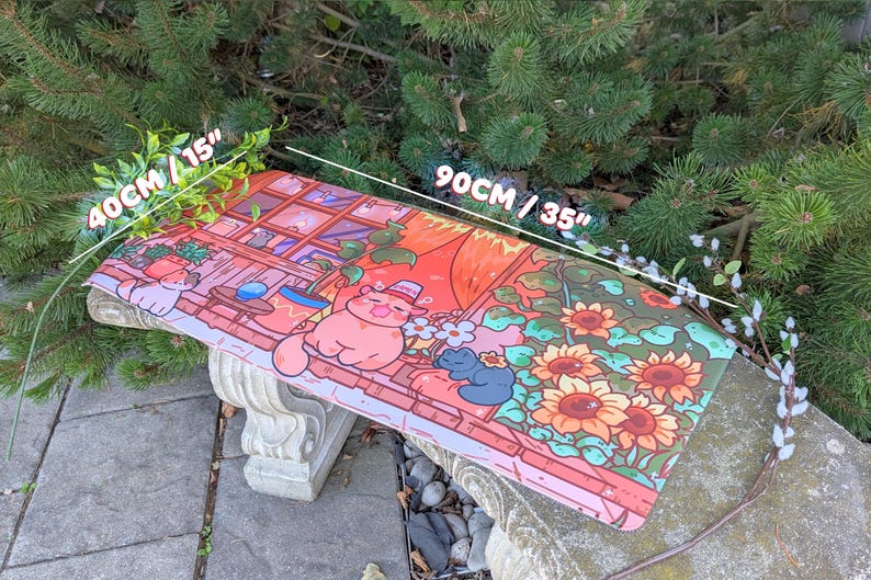 May include: A rectangular desk mat with a colorful illustration of a cat in a hat, sunflowers, and a window scene. The mat measures 35 inches long and 15 inches wide. The design features shades of pink, orange, and green.