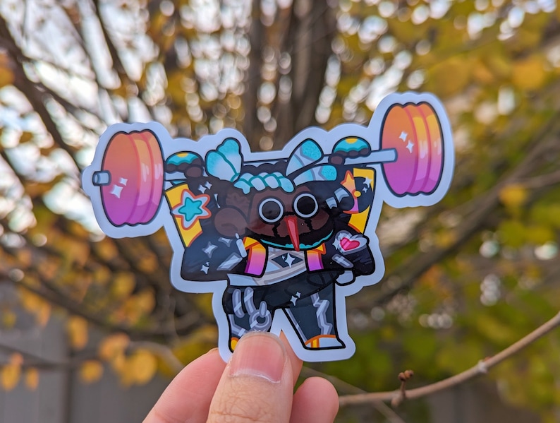 May include: A colorful sticker of a cartoon character lifting a pink and orange barbell. The character has black eyes, a black nose, and a pink heart on its chest. The character is wearing a black and white outfit with a blue and white design on its head.