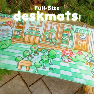 May include: A rectangular deskmat featuring a colorful illustration of a frog-themed cafe with green checkered floors, a pastry display case, tables, and chairs. The text "Full-Size deskmats!" is printed in white at the top of the mat.