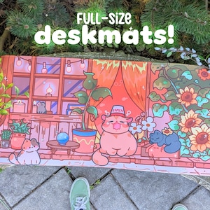 May include: Full-size deskmat featuring a whimsical illustration of a cat in a hat, surrounded by plants, flowers, and other cats. The desk mat has a warm color palette with a window scene and the text "FULL-SIZE deskmats!"