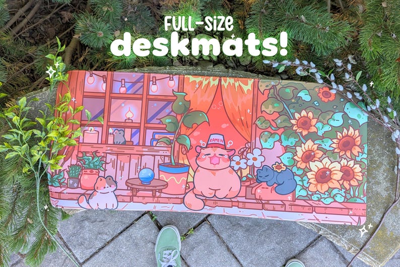 May include: Full-size deskmat featuring a whimsical illustration of a cat in a hat, surrounded by plants, flowers, and other cats. The desk mat has a warm color palette with a window scene and the text "FULL-SIZE deskmats!"