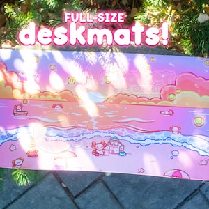 May include: A rectangular deskmat featuring a vibrant pink and orange sunset beach scene with cartoon axolotls, a sandcastle, and floating lanterns. The text "FULL-SIZE deskmats!" is printed in white and pink at the top of the illustrated surface.