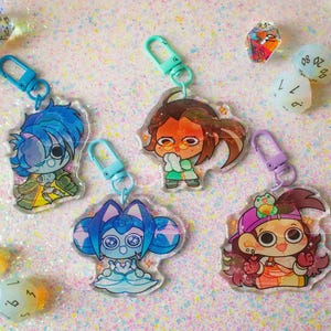 May include: Four acrylic keychains featuring chibi-style anime characters with colorful designs, each attached to a plastic lobster clasp in blue, mint green, or purple, surrounded by several multi-faceted polyhedral dice on a glittery surface.