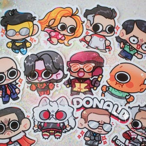 Puede incluir: Una colección de once pegatinas de vinilo troqueladas con personajes estilo chibi de ojos grandes. Los diseños incluyen varias figuras con trajes de superhéroes y ropa formal, con una pegatina que muestra el texto DONALD en letras negritas.