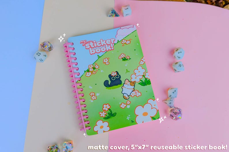 May include: A reusable sticker book with a matte cover, measuring 12.7cm x 17.8cm. The cover shows a green field with cartoon cats and flowers. The book has a pink spiral binding. Dice are scattered around the book.