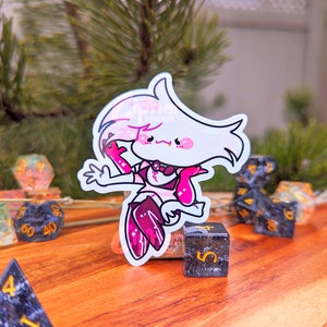 May include: A pink and white cartoon character sticker with a pink bow and a pink tongue sticking out. The sticker is on a wooden surface with black and gold dice.