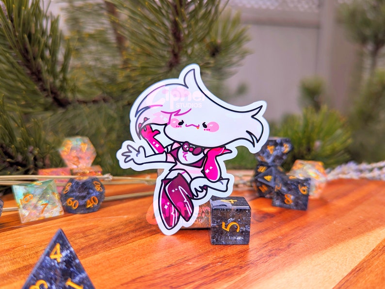May include: A pink and white cartoon character sticker with a pink bow and a pink tongue sticking out. The sticker is on a wooden surface with black and gold dice.
