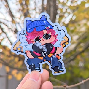 May include: A cartoon character sticker with a blue beanie, pink hair, and white fur. The character is wearing a black and blue outfit and has a pink heart on their beanie. The sticker is on a blurry background of fall foliage.