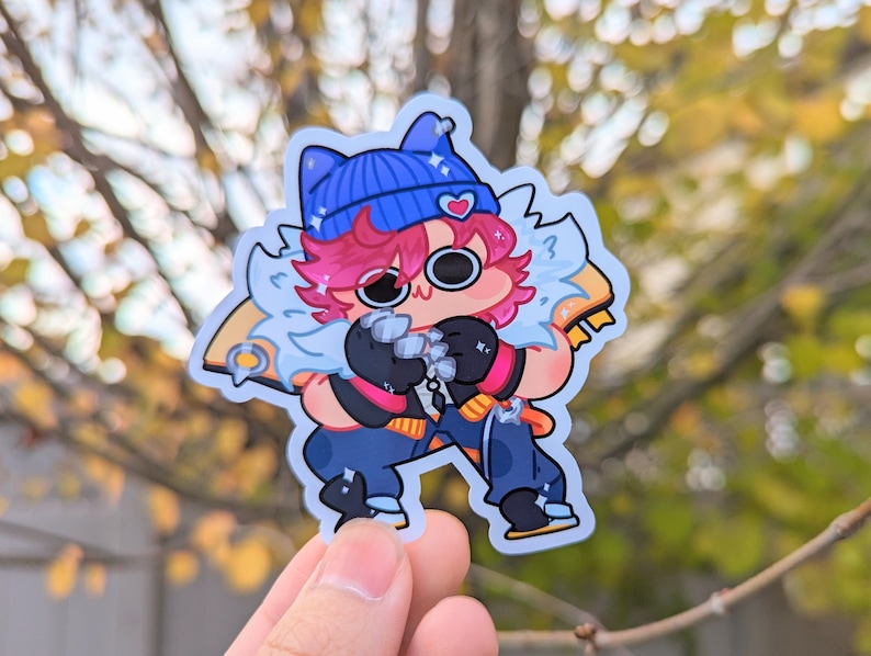 May include: A cartoon character sticker with a blue beanie, pink hair, and white fur. The character is wearing a black and blue outfit and has a pink heart on their beanie. The sticker is on a blurry background of fall foliage.