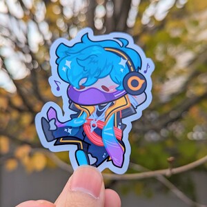 May include: A blue and purple cartoon character sticker with a yellow headband and headphones. The character is wearing a purple mask and has blue hair. The sticker is on a white background.