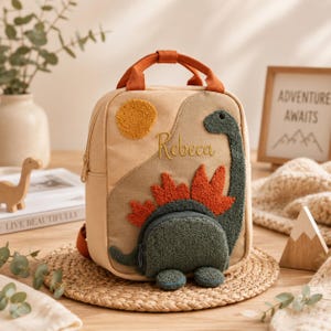 May include: A beige canvas backpack featuring a textured, dark green dinosaur appliqué with orange spikes, a yellow sun, and the embroidered name Rebeca. The bag includes orange top handles, a front zippered pocket, and gold-toned hardware.