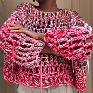 May include: A crocheted sweater with a loose, open weave in shades of pink, red, and white. The sweater has a relaxed fit and a slightly cropped length.