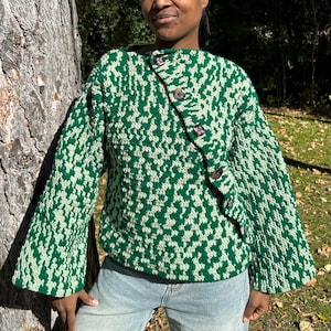 Könnte beinhalten: Ein grün-weiß gemusterter Pullover mit einem einzigartigen Knopfverschluss. Der Pullover hat lange Ärmel und ist aus einem strukturierten Strickstoff gefertigt. Das Model trägt eine blaue Jeans und steht draußen in der Nähe eines Baumes.