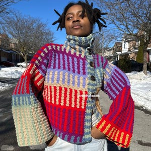 Puede incluir: Una chaqueta de crochet colorida con cuello alto y mangas acampanadas. La chaqueta está hecha en estilo patchwork con rayas rojas, moradas, azules y crema.