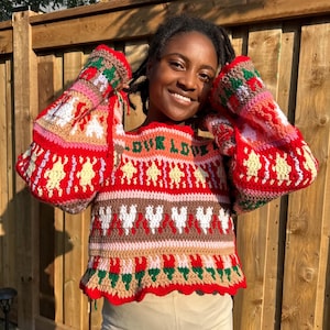 May include: A red, white, and brown crocheted sweater with a ruffled hem. The sweater has a pattern of hearts, stars, and flowers. The text "LOVE LOVE" is written on the front of the sweater.