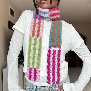 May include: A colorful, striped crochet scarf in shades of pink, green, blue, and brown. The scarf is handmade with a textured stitch and features vertical stripes. The text "crochetaco" is visible on the scarf.