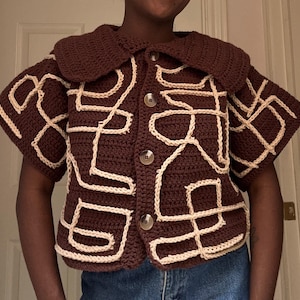 May include: A brown crocheted cardigan with a large white crocheted collar and short sleeves. The cardigan has a geometric pattern of white lines on a brown background.