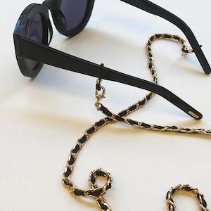 May include: Black sunglasses with a gold chain attached to the arms. The chain is made of a braided black cord with gold links.