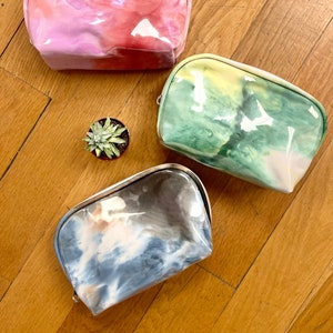 May include: Three colorful, translucent cosmetic bags with watercolor-like designs. One is pink and red, another is green and yellow, and the third is blue and gray. A small succulent plant sits between the bags.