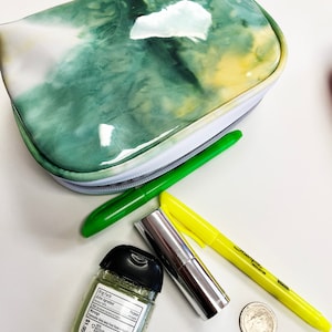 Könnte beinhalten: Eine grün-gelbe Tie-Dye-Schminktasche mit Reißverschluss. Die Tasche ist geöffnet und enthält einen grünen Stift, einen gelben Textmarker, eine silberne Lippenstifttube und eine durchsichtige Plastikflasche mit Handdesinfektionsmittel und schwarzem Deckel. Das Etikett der Flasche lautet "Stay Fresh" und enthält eine Liste der Inhaltsstoffe und Warnhinweise. Die Tasche liegt auf einer weißen Oberfläche.
