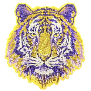 May include: A sequined tiger head patch in gold, purple, and white. The tiger's face is centered, with yellow sequins outlining the shape. The eyes are yellow, and the stripes are purple. The patch is likely for clothing or accessories.