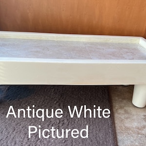 May include: Antique white wooden dog bed with a raised platform and four legs. The bed is pictured on a brown rug. "Antique White Pictured" is written in black text below the bed.
