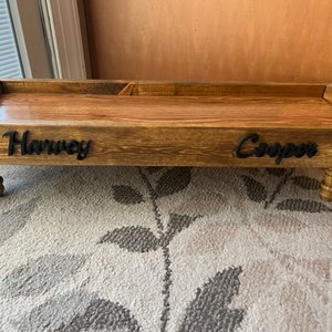 May include: A wooden dog bed with raised edges and four legs. The bed is stained a dark brown colour and has the names "Harvey" and "Cooper" engraved on the front.