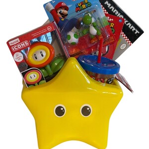 K&ouml;nnte beinhalten: Ein gelber sternf&ouml;rmiger Beh&auml;lter gef&uuml;llt mit Spielzeug und Waren zum Thema Super Mario. Enth&auml;lt eine Mario-Figur, eine Yoshi-Figur, ein Mario Kart-Spiel und ein Paladone Icons-Spielzeug. Ein blau-roter Becher mit Strohhalm ist ebenfalls sichtbar.