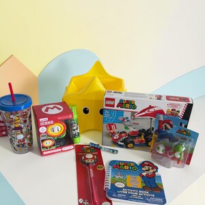K&ouml;nnte beinhalten: Eine Sammlung von Super Mario-Artikeln, darunter ein Becher, ein Lego-Set, ein Stift, ein Aktivit&auml;tsbuch und Figuren. Die Artikel sind auf einer wei&szlig;en Oberfl&auml;che mit einem gelben sternf&ouml;rmigen Beh&auml;lter im Hintergrund angeordnet.