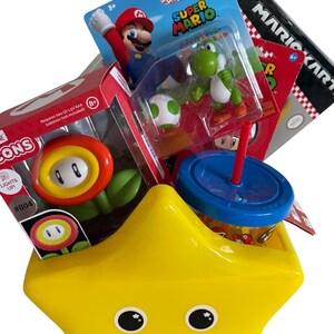 K&ouml;nnte beinhalten: Ein gelber sternf&ouml;rmiger Beh&auml;lter gef&uuml;llt mit Super Mario Spielzeug und Merchandise. Die Artikel umfassen Actionfiguren von Mario und Yoshi, ein Feuerblumen-Spielzeug, ein Mario Kart-Spiel und einen blau-roten Becher mit Strohhalm.