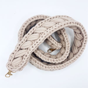 May include: A beige, braided strap with gold-colored metal clasps. The strap is made of a soft, woven material and is designed to be used with a bag or purse. The strap is shown against a white background.