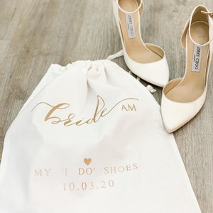 Peut inclure: Un sac en toile blanche à cordon avec le texte "Bride AM" en écriture dorée. Le sac porte également le texte "My 'I Do' Shoes 10.03.20" en lettres dorées. Deux chaussures blanches à talons hauts avec des boucles dorées sont posées sur le sac.