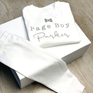 Op de afbeelding: Witte baby outfit met een shirt met lange mouwen en een broek. Het shirt heeft een grijze strik en de tekst "Page Boy Parker" erop gedrukt.