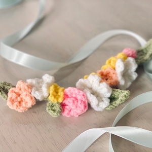 May include: A crocheted flower garland with pink, white, yellow, and green flowers. The garland is on a light blue ribbon.
