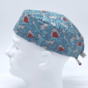 May include: A scrub cap featuring a repeating pattern of grey sharks with open mouths and red interiors swimming through stylized blue waves. The cap includes a small white fabric tag and a cord with a red bead at the back for adjustment.