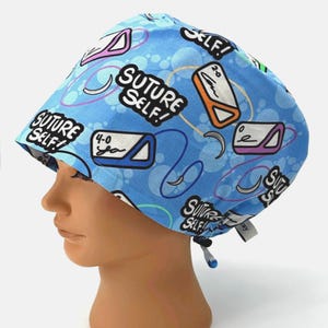 May include: A light blue surgical scrub cap with a repeating pattern of medical illustrations and the text "Suture Self!". The cap features a drawstring for an adjustable fit.