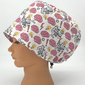 May include: White scrub cap with a repeating pattern of pink brains, yellow light bulbs, red exclamation points, and lightning bolts. The cap has the text "Hold On, You're Overthinking This". The cap has a black drawstring.