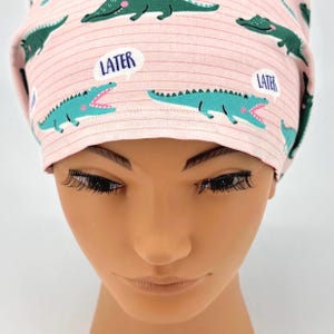 May include: A pink scrub cap with a repeating pattern of teal alligators and speech bubbles that say "LATER". The cap has a striped background.