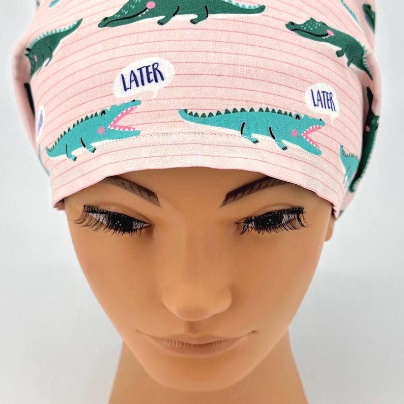 May include: A pink scrub cap with a repeating pattern of teal alligators and speech bubbles that say "LATER". The cap has a striped background.