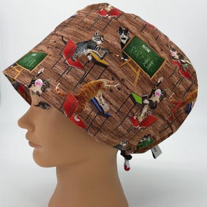 May include: A brown and tan scrub hat with a repeating pattern of cats in a classroom setting. The cats are wearing glasses and holding books. The text "MEOW" is written on a chalkboard.