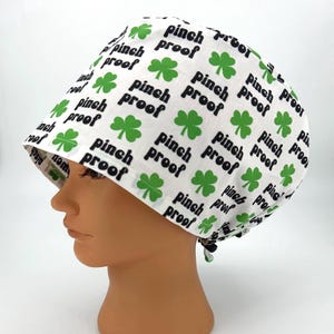 May include: White surgical scrub cap with a repeating pattern of green shamrocks and the words "pinch proof" in black. The cap is tied in the back and is designed to cover the hair.
