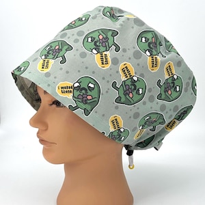 May include: A light green surgical scrub cap featuring a repeating pattern of cartoon zombie characters holding ice pops. Each zombie has a sad expression and the text "I made these" is in a speech bubble. The cap has a drawstring.