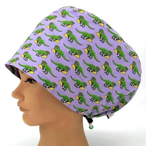 May include: A lavender surgical scrub cap featuring a repeating pattern of green dinosaurs and yellow tacos. The cap is designed with a comfortable fit and a drawstring for adjustment. This cap is suitable for medical professionals.