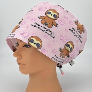 May include: A light pink scrub cap featuring a repeating pattern of cartoon sloths in a yoga pose, spiral designs, and stars. The cap has the text "Relax, everything is perfectly out of control!"