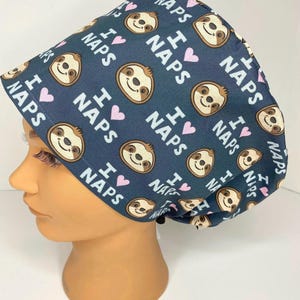May include: A navy blue surgical scrub cap featuring a repeating pattern of smiling sloths, pink hearts, and the text "I ❤️ NAPS" in white. The cap is designed to cover the head and hair.
